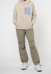 Fleece hoodie in lichtbeige met een blauwe borstzak, gecombineerd met groene broek. Voorzien van een ritssluiting aan de voorkant, een trekkoord in de zoom en witte sneakers.