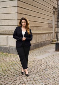 Blazer nero su top bianco, pantaloni neri e tacchi neri. Modella in piedi su una strada di ciottoli vicino a un muro di pietra testurizzato.