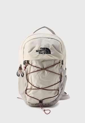 Jasnobeżowy mini plecak The North Face Borealis z elastycznym sznurkiem z przodu i dwoma zapinanymi na suwak kieszeniami, przedstawiony w pozycji pionowej na białym tle.