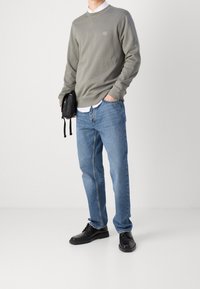 Man som bär en grå sweatshirt över en vit skjorta, blå jeans, svarta läderskor, och håller en svart påse mot en plain vit bakgrund.