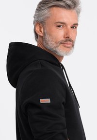 Sudadera negra con corte regular, con capucha ajustable, puños de punto acanalado y un parche con logo en la manga izquierda. Tejido suave de mezcla de algodón.
