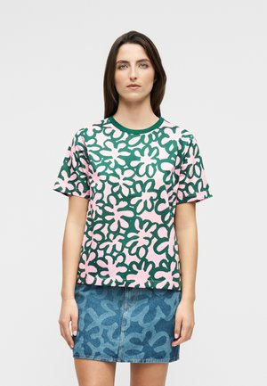 Kvinde med glat brunt hår iført en grøn og pink kortærmet skjorte med blomsterprint samt et blåt mønstret denimskørt.