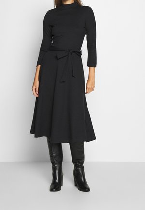 Femme portant une robe midi noire avec une ceinture à la taille, des manches 3/4 et des bottes noires à talons montantes, se tenant devant un fond uni.