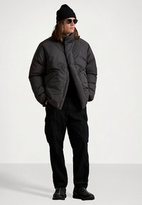 Tumši pelēka pūdera jaka ar augstu apkakli un priekšējo rāvējslēdzu. Kopā ar melnām darba biksēm un izturīgiem melniem apaviem. Aksesuāri: melna adīta cepure un saulesbrilles.