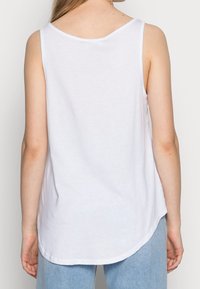 Top blanco sin mangas con escote redondeado, hecho de tela suave; cuenta con un dobladillo curvado y un ajuste relajado, combinado con jeans de un azul claro.