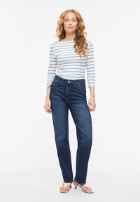 Γυναίκα με μακριά σγουρά ξανθά μαλλιά, φορώντας λευκό και μπλε ριγωτό τοπ, σκούρο μπλε τζιν και λευκά σανδάλια με τακούνι, που στέκεται σε λευκό φόντο.