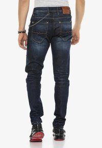 Dunkelblaue Slim-Fit-Jeans mit ausgewaschenen Details, fünf Taschen, orangefarbenen Nähten und einem braunen Lederpatch, getragen mit schwarzen, mit Nieten besetzten Sneakers.