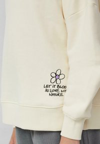 Felpa color crema con polsini a coste. Presenta un ricamo con un design floreale e la scritta "LASCIALO FIORIRE NELL'AMORE PER LA NATURA" sotto.