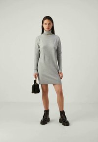 Pilkas megztas turtlenecko suknelė su ilgomis rankovėmis, turinti lygų apdailą. Suderinta su juodais šnuoruojamais batais ir mažomis tekstūruotomis rankinėmis.