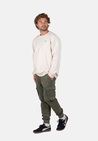 Sudadera beige claro, pantalones cargo oliva con bolsillos laterales y zapatillas negras con detalles en gris y blanco. El modelo está de pie sobre un fondo liso.