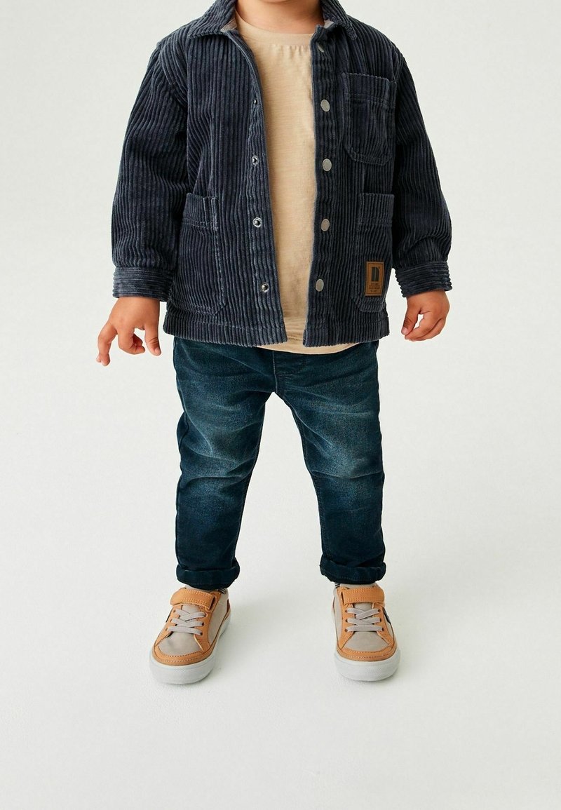 Tout-petit porte une chemise beige, une veste en velours côtelé bleu foncé avec des boutons, un jean bleu foncé et des baskets beige et marron clair, debout sur fond blanc.