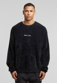Jersey negro de pelo suave con mangas largas, escote redondo y texto bordado "Otro Algodón" en el pecho. Textura suave, corte relajado.