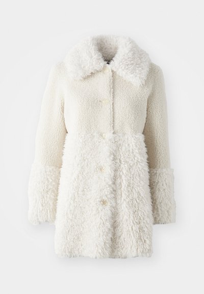 Valge karvane mantel, millel on tekstureeritud kangas, suur krae ja nööbid. Keha on sileda viimistlusega, kontrastsete aktsentidega.