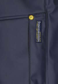 Marinblått vattentåligt tyg med en sydd söm och en gul logoknapp. Inkluderar en tygetikett med vit och gul text.