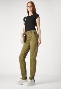 Alyvuogių spalvos kelnės su siaurėjančiu dizainu, su šoninėmis užtrauktukų kišenėmis ir priekinėmis kišenėmis. Sujungtos su juoda prigludusia marškinėliais ir baltais sportiniais bateliais.