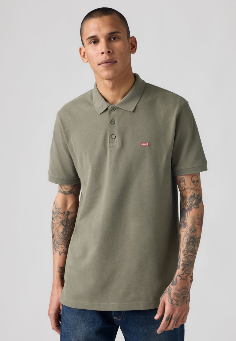 Homme aux cheveux courts portant un polo Levi's vert olive, montrant des bras entièrement tatoués, debout devant un fond gris clair uni.
