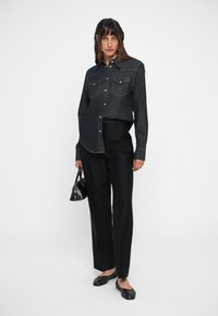 Donkerdenim overhemd met een knoopsluiting, voorzien van twee borstzakken en gele stiksels, gecombineerd met zwarte op maat gemaakte broek en schoenen.