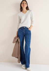 Lichtgrijze trui met een V-hals, gecombineerd met donkerblauwe flared jeans. Het model houdt een beige jas vast en draagt witte sneakers. Over het geheel is er een gladde textuur.