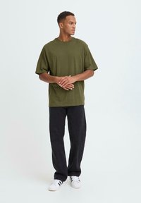 Tricou peste mărime din bumbac, de culoare verde măsliniu, cu mâneci scurte, asortat cu pantaloni negri largi și adidași albi. O ținută simplă și casual.