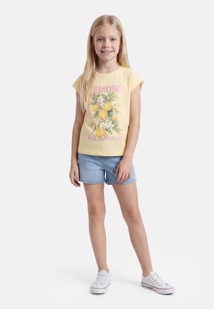 Ung pige med langt blondt hår iført gul t-shirt med citronprint, denimshorts og hvide sneakers, der står og smiler.