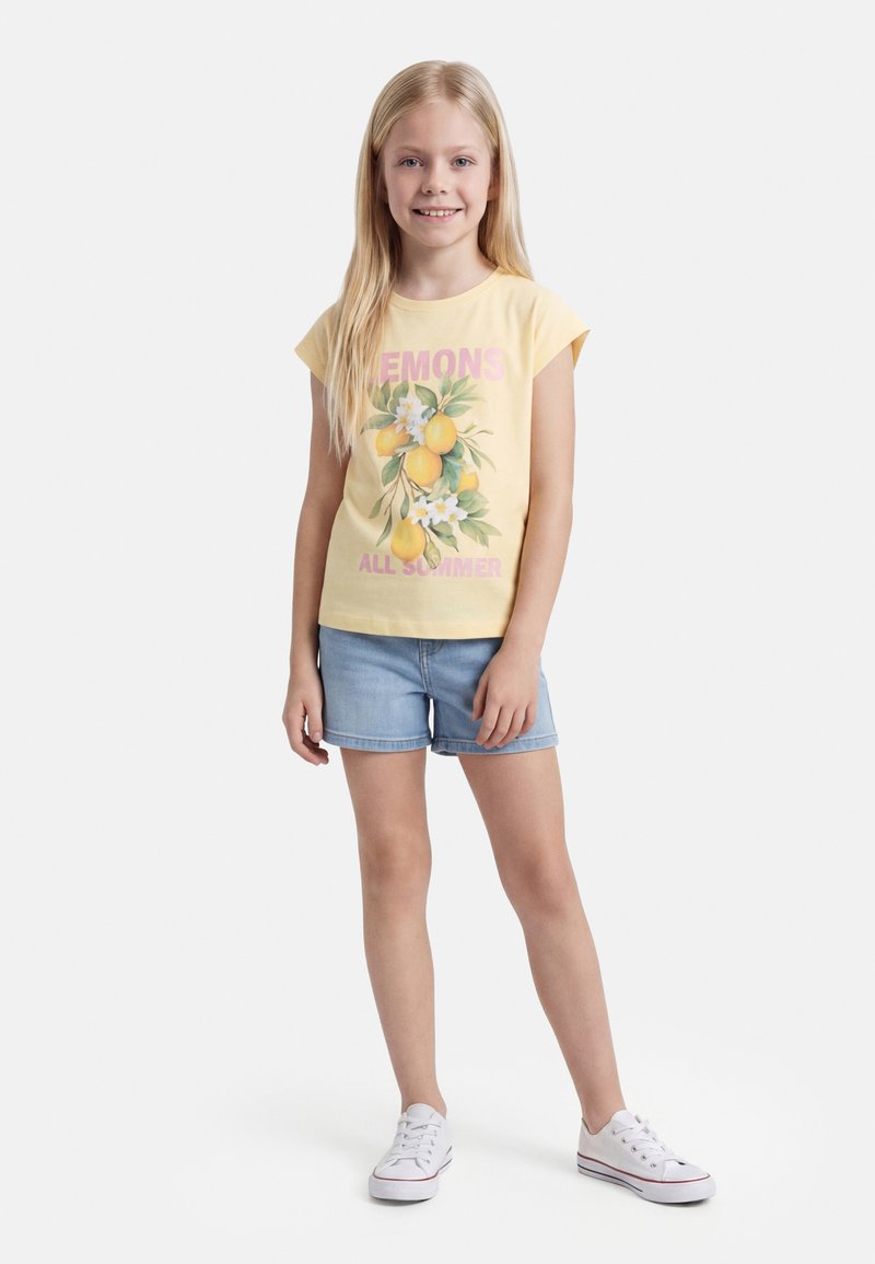 Νεαρό κορίτσι με μακριά ξανθά μαλλιά, που φορά κίτρινο μπλουζάκι με τύπωμα λεμονιού, τζιν σορτς και λευκά αθλητικά παπούτσια, στέκεται και χαμογελά.