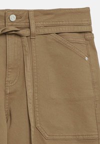 Gros plan d'un pantalon beige avec une ceinture en tissu, un bouton en métal, une poche avant et des détails de couture visibles sur un tissu en coton.