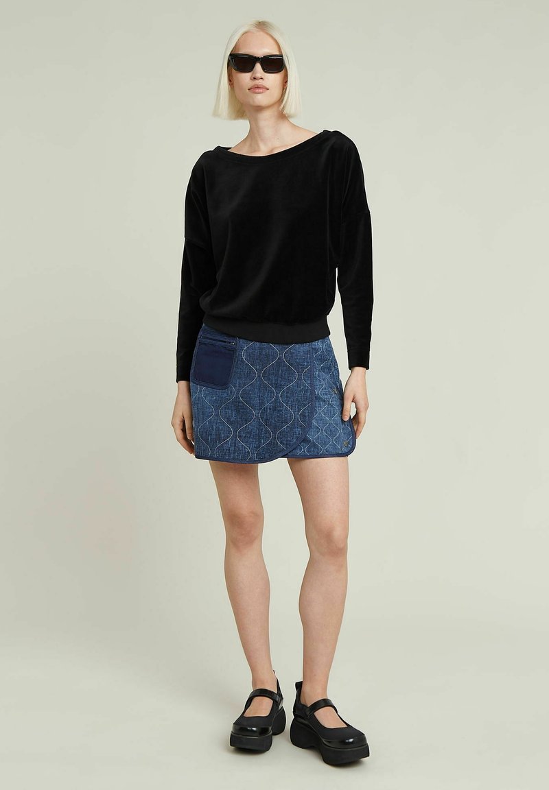 Schwarzes Samtoberteil mit langen Ärmeln, kombiniert mit einem blauen Jeansrock mit Wellenmustern und einer aufgesetzten Tasche. Schwarze Plateauschuhe runden das Outfit ab.