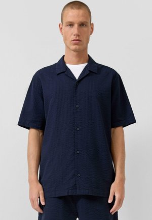 Mężczyzna z krótkimi włosami, ubrany w granatową, teksturowaną koszulę z krótkim rękawem i guzikiem, na białej koszulce z okrągłym dekoltem, na jednolitym jasnym tle.