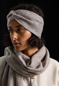Femme à la peau brune et aux cheveux courts et bouclés, portant un bandeau en tricot gris et une écharpe assortie, se tournant légèrement sur le côté devant un fond noir.