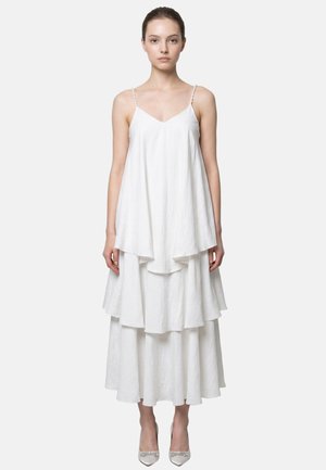 Donna che indossa un vestito bianco senza maniche a balze con spalline sottili e décolleté bianche a punta, in piedi contro uno sfondo semplice.