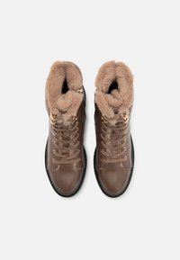 Bottines montantes en cuir marron avec une doublure en fourrure douce, œillets dorés pour les lacets et des accents texturés au niveau de la cheville. Semelle en caoutchouc noire.