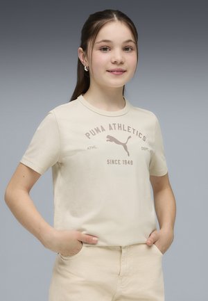 Joven con cabello oscuro, vestido con camiseta y pantalones color crema de Puma Athletics, de pie con las manos en los bolsillos contra un fondo gris liso.