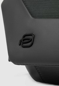 Schwarzes, strukturiertes Material mit geometrischer Form, das ein glänzendes schwarzes Logo deutlich zeigt. Dezente Muster erhöhen die visuelle Attraktivität.