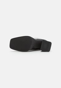 Suela de zapato negra con una superficie de goma texturizada, con puntera cuadrada y un tacón firme. Acento sobresaliente con el nombre de la marca grabado.