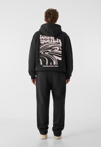 Crna dukserica s velikim, apstraktnim ružičastim uzorkom na leđima, koja sadrži tekst "izgubljena mladost" i "Kreativnost se nalazi u haosu." Uparen s crnim trenerkama.