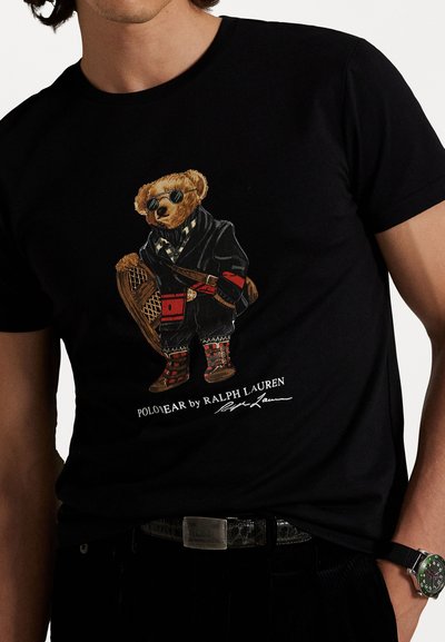 Melns kokvilnas t-krekls ar lāča grafiku, kas tērpies mētelī, valkā saulesbrilles un rakstainus zābakus. Apakšā ieraksts "POLO BEAR by RALPH LAUREN".