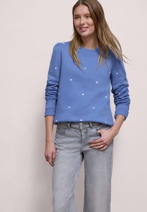 Femme aux longs cheveux portant un sweat-shirt bleu à motifs de cœurs blancs et un jean gris clair, souriante avec la main dans la poche.