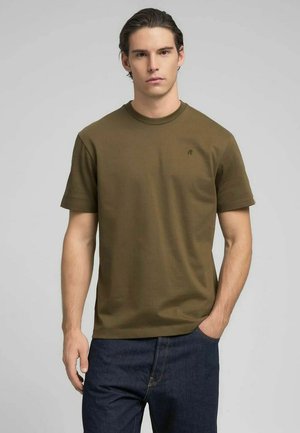 T-shirt a maniche corte verde oliva in tessuto a coste con scollo rotondo, caratterizzata da un piccolo logo sobrio sul petto. Abbinata a jeans scuri.