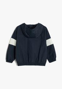Giacca a vento blu navy con cappuccio, caratterizzata da strisce diagonali bianche sulle maniche e orlo ed elastici alle maniche.