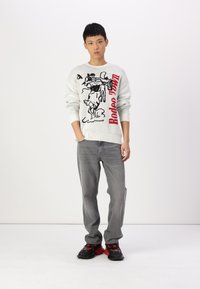 Witte gebreide trui met een rodeo-graphic en rode tekst, gecombineerd met grijze jeans en zwarte sneakers met rode accenten.