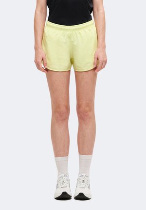 Person iført lysegule shorts, hvide ankelsokker, hvide sneakers og en sort skjorte, der står mod en ensfarvet baggrund.