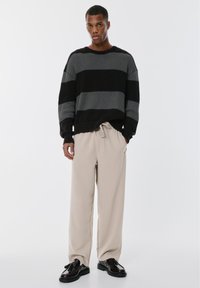 Crna i siva prugasta pletena dukserica, opuštenog kroja, uparena s bež plisiranim hlačama i tamnim cipelama, prikazuje glatku tkaninu i krojeni dizajn.