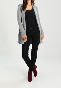 Cardigan lungo grigio a coste indossato sopra una maglia nera, abbinato a jeans neri e stivali ankle bordeaux. Texture morbide con una vestibilità rilassata.