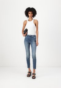 Top a righe bianco, jeans attillati blu, borsa nera a tracolla e sandali neri con cinturini e punta aperta. Sfondo neutro.