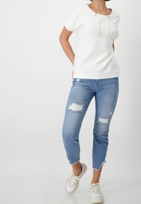 Valkoinen lyhythihainen paita, jossa on kiristettävä kaula-aukko, yhdistettynä vaaleansinisiin kulutettuihin farkkuihin, joissa on koristeita ja repsottavat lahkeet, sekä valkoiset lenkkarit.