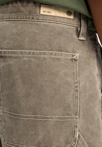 Pantaloni cargo in tessuto marrone chiaro con tasca applicata e cuciture a contrasto. L'etichetta sulla vita reca la scritta "EST. 1992." Materiale testurizzato e resistente.