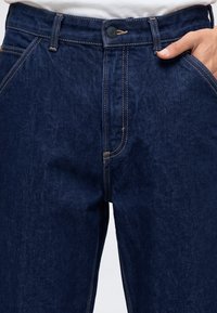 Σκούρα μπλε τζιν denim με αντίθετες ραφές, κλείσιμο με κουμπί, θηλιές για ζώνη και ένα χέρι τοποθετημένο στην μπροστινή δεξιά τσέπη.