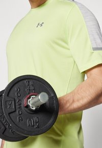 Osoba ubrana w limonkową koszulkę Under Armour podnosi czarny hantel 2 kg prawą ręką zgiętą w łokciu.