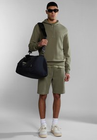 Sudadera verde oliva con cordón, pantalones cortos a juego, zapatillas blancas y bolsa de deporte negra con asas laterales y logo. Fondo gris.