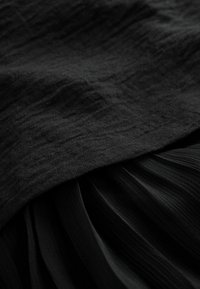 Černá tkanina s texturovanou, zvrásněnou vrchní vrstvou nad hladkými, skládacími vrstvami. Celkový design je monochromatický s různými texturami.